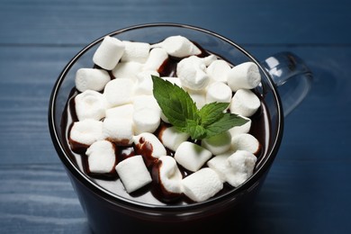 Photo of Glass cup of delicious hot chocolate with marshmallows and fresh mint on blue wooden table, closeup