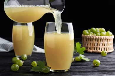 Photo of Pouring tasty gooseberry juice from jug into glass on black wooden table against dark background