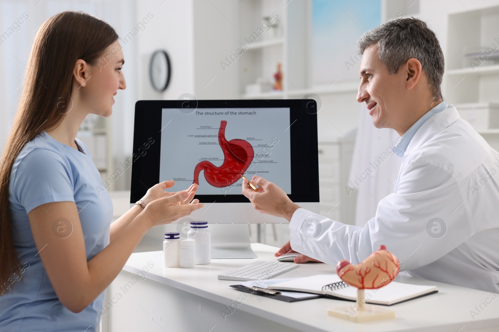 Photo of Gastroenterologist showing screen with illustration of human stomach model to patient at table in clinic