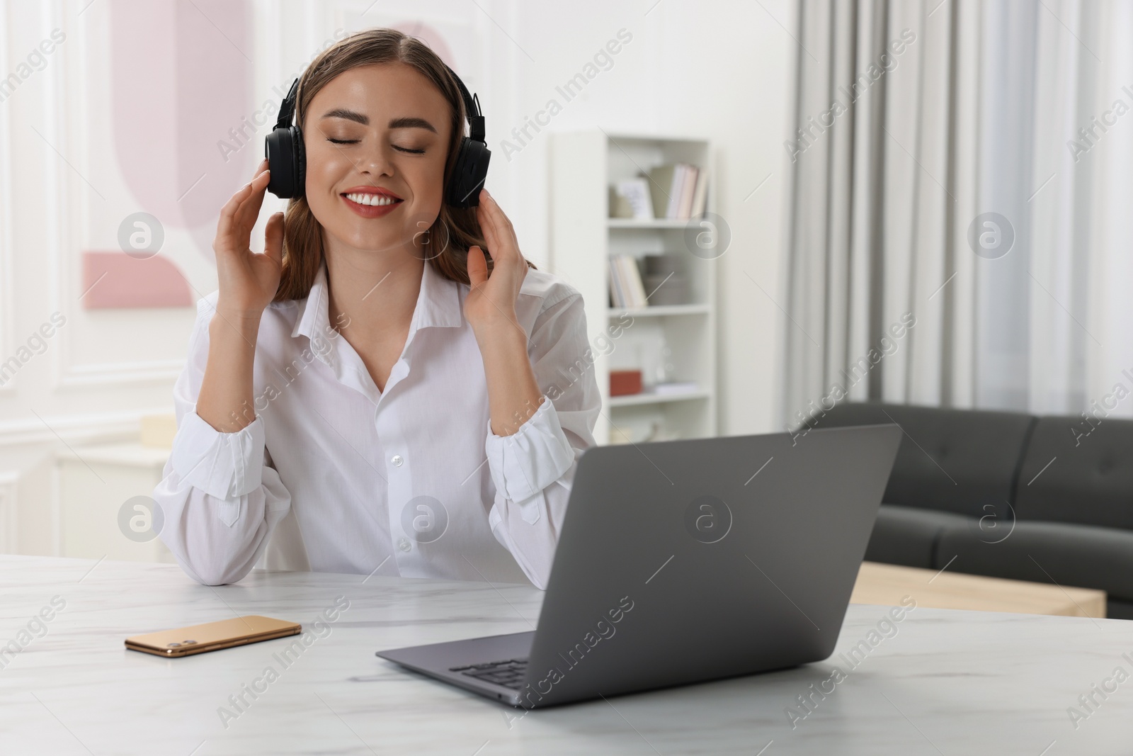Photo of Happy woman with headphones listening to music near laptop at white table in room