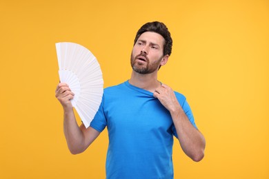 Photo of Unhappy man with hand fan suffering from heat on orange background