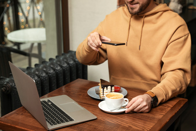 Photo of Male blogger taking photo of dessert and coffee at table in cafe, closeup