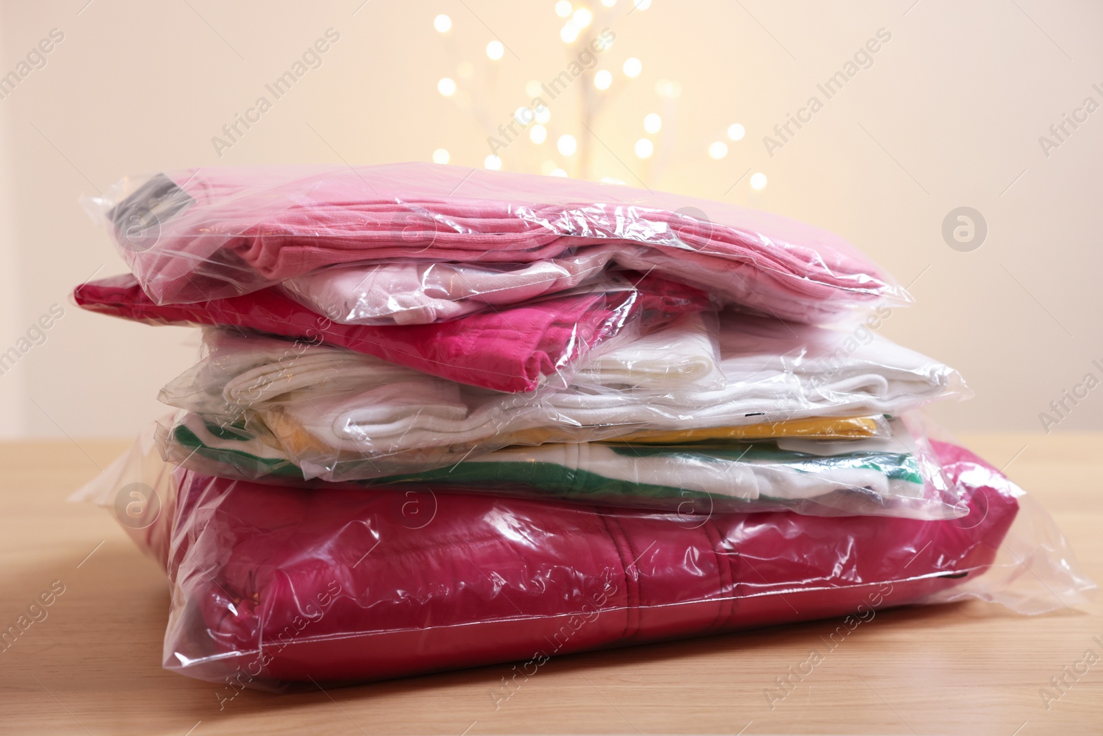 Stack of folded clothes in plastic bags on wooden table against beige background with blurred lights Photo of Stack of folded clothes in plastic bags on wooden table against beige background with blurred lights