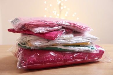 Photo of Stack of folded clothes in plastic bags on wooden table against beige background with blurred lights