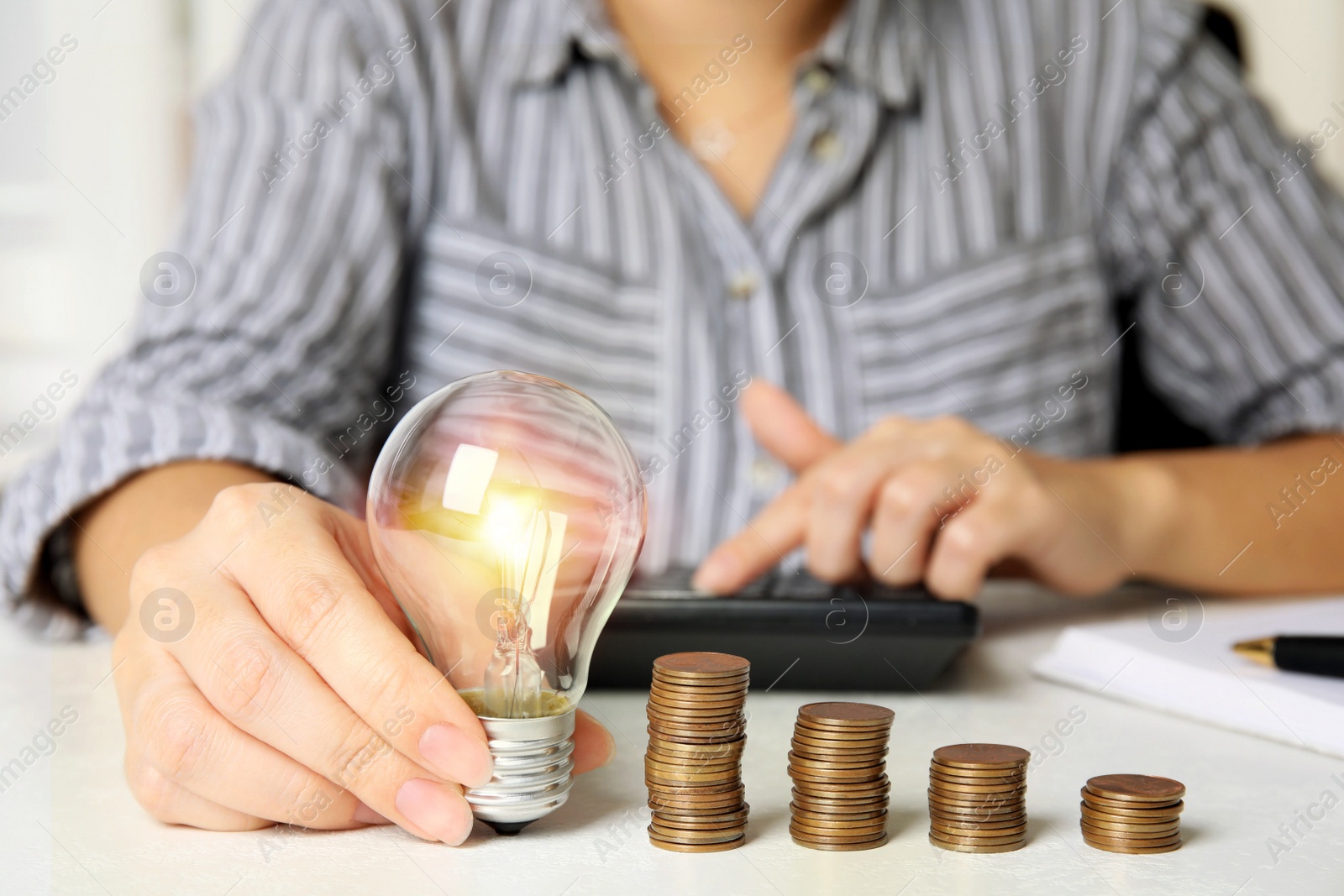 Photo of Woman with light bulb, calculator and coins at white table, closeup. Power saving