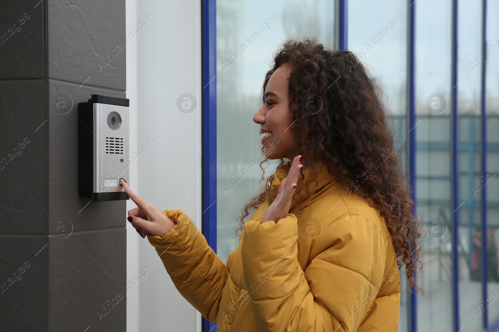 Photo of Young African-American woman ringing intercom while waving to camera near building entrance