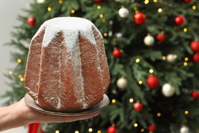 Photo of Woman holding delicious Pandoro cake decorated with powdered sugar near Christmas tree, closeup and space for text. Traditional Italian pastry