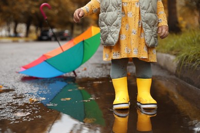 Photo of Little girl wearing yellow rubber boots standing in puddle outdoors, closeup. Space for text