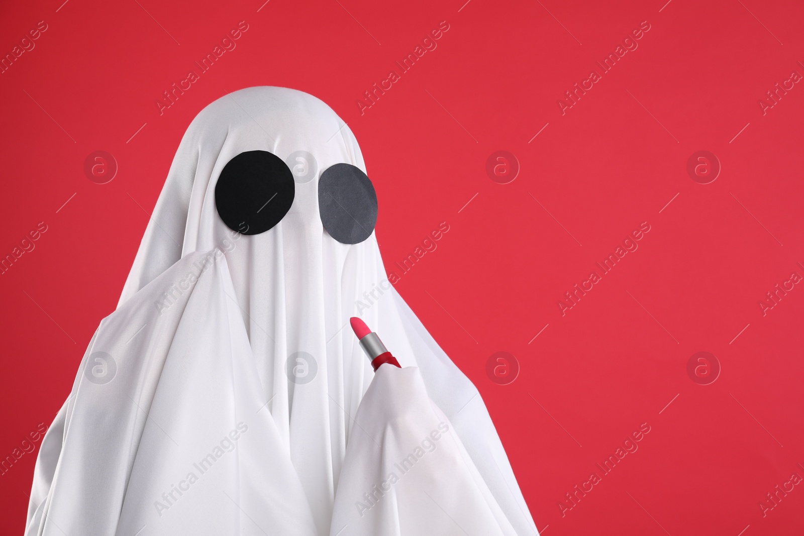 Photo of Fashionable ghost. Woman covered with white sheet applying lipstick on red background, space for text