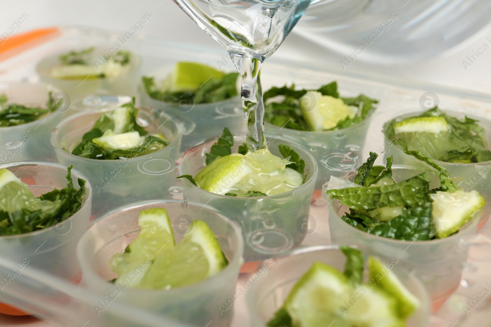 Photo of Pouring water into ice cube tray with lime slices and mint, closeup