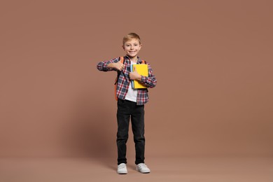 Photo of Happy schoolboy with backpack and books showing thumb up gesture on brown background