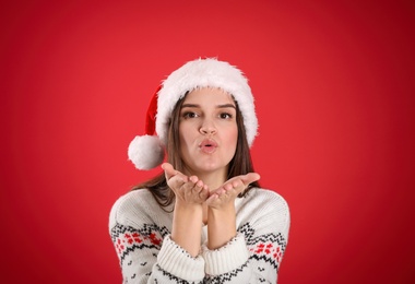 Photo of Pretty woman in Santa hat and Christmas sweater blowing kiss on red background