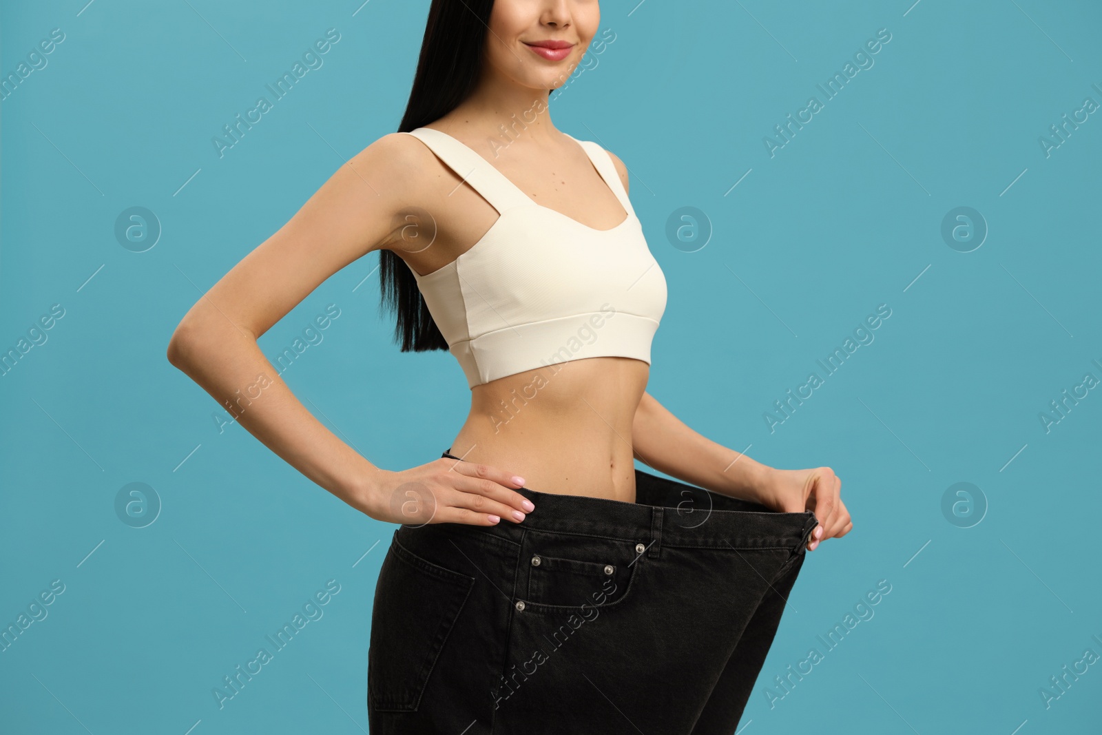 Photo of Young woman wearing big jeans after weight loss on turquoise background, closeup