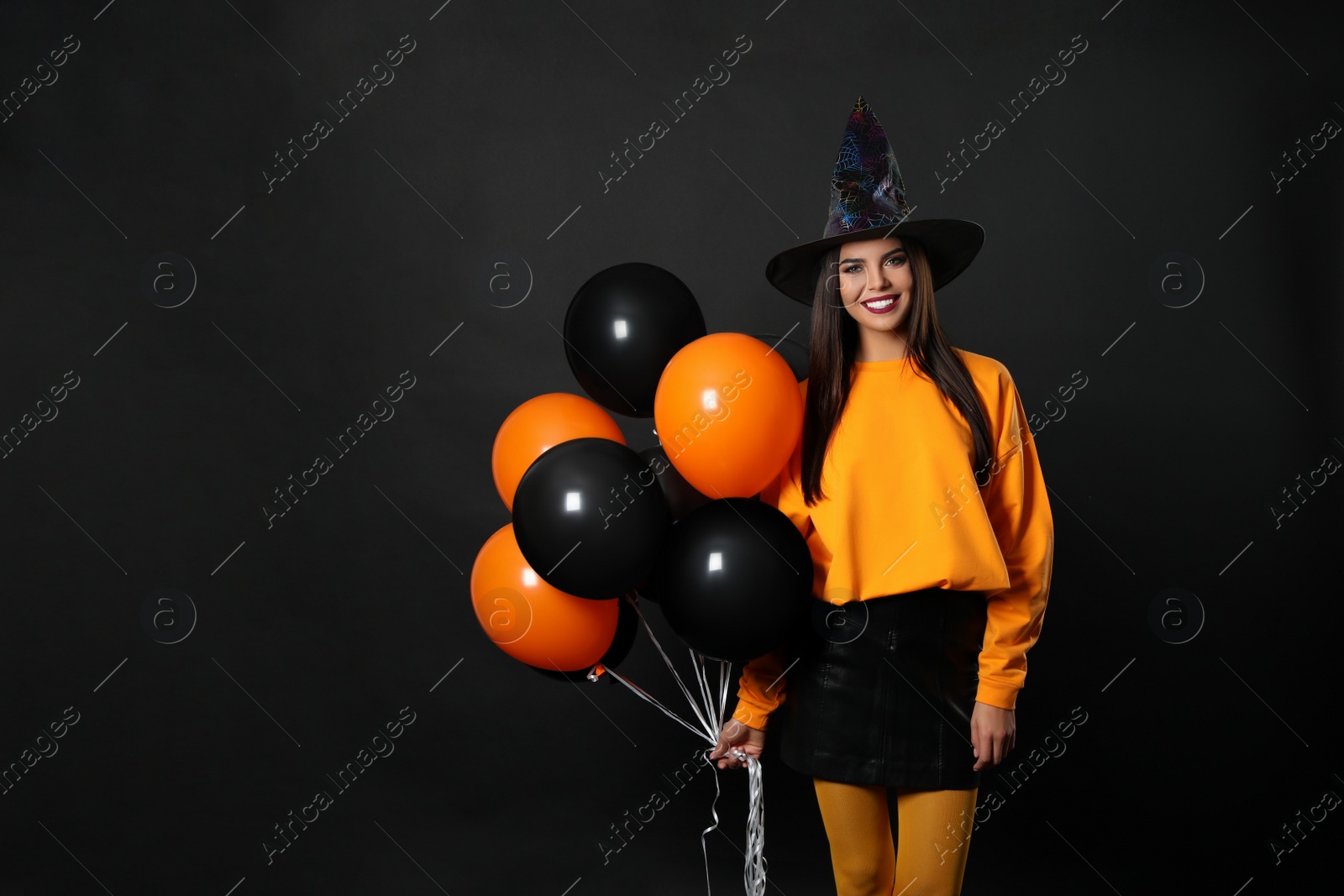 Photo of Beautiful woman wearing witch costume with balloons for Halloween party on black background, space for text