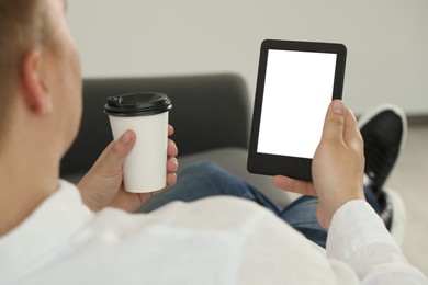 Man with cup of coffee using e-book reader indoors, closeup