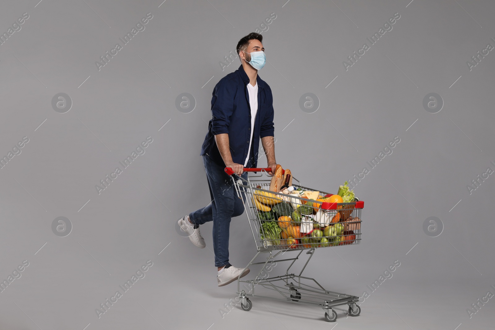 Photo of Man with protective mask and shopping cart full of groceries on light grey background