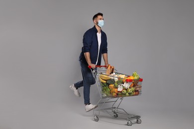 Man with protective mask and shopping cart full of groceries on light grey background