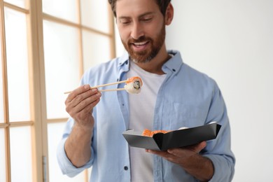 Photo of Happy man eating sushi rolls with chopsticks indoors, focus on hand