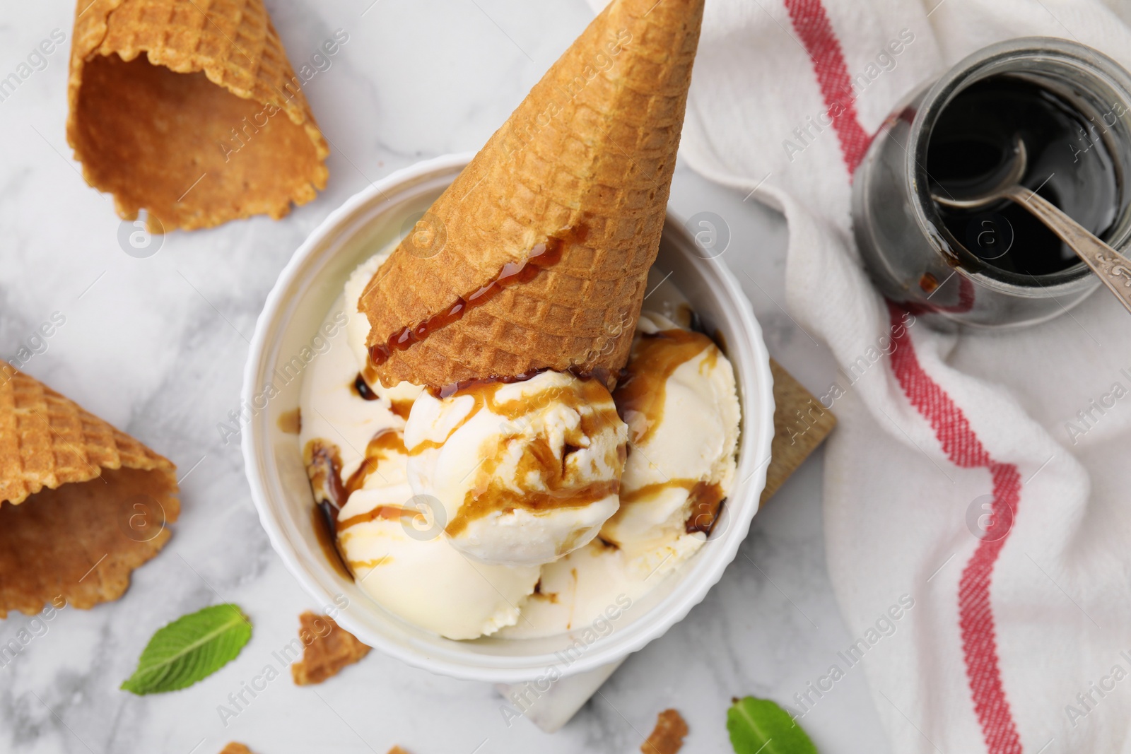 Photo of Scoops of ice cream with caramel sauce, mint leaves and cone on white marble table, flat lay