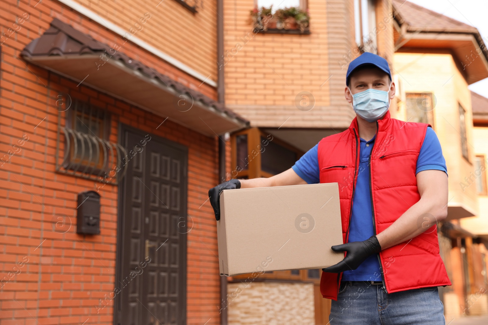 Photo of Courier in protective mask and gloves with box near house outdoors. Delivery service during coronavirus quarantine