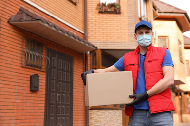 Courier in protective mask and gloves with box near house outdoors. Delivery service during coronavirus quarantine