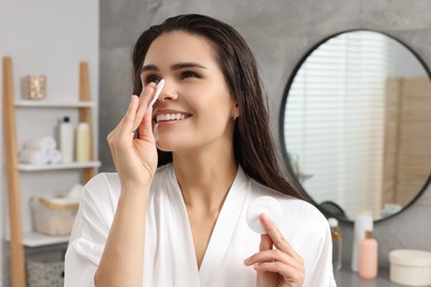 Young woman cleaning her face with cotton pad in bathroom