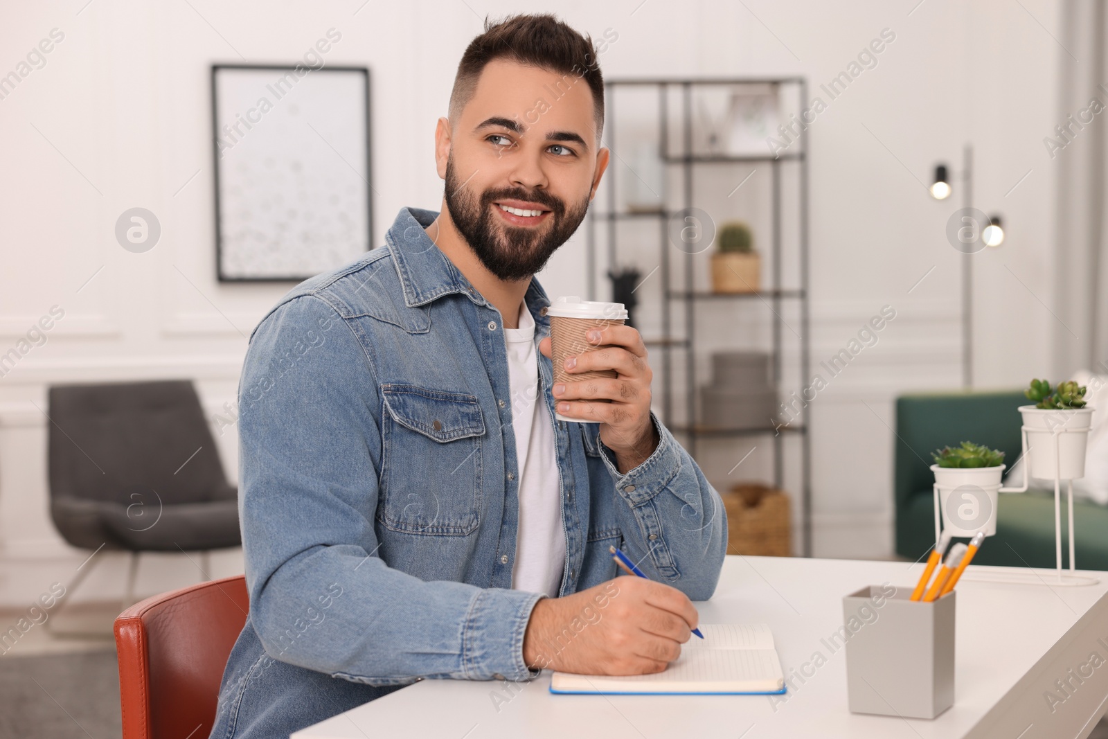 Photo of Young man with cup of coffee writing in notebook at white table indoors