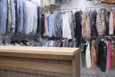 Dry-cleaning service. Hangers with different clothes in plastic bags on racks and wooden counter indoors