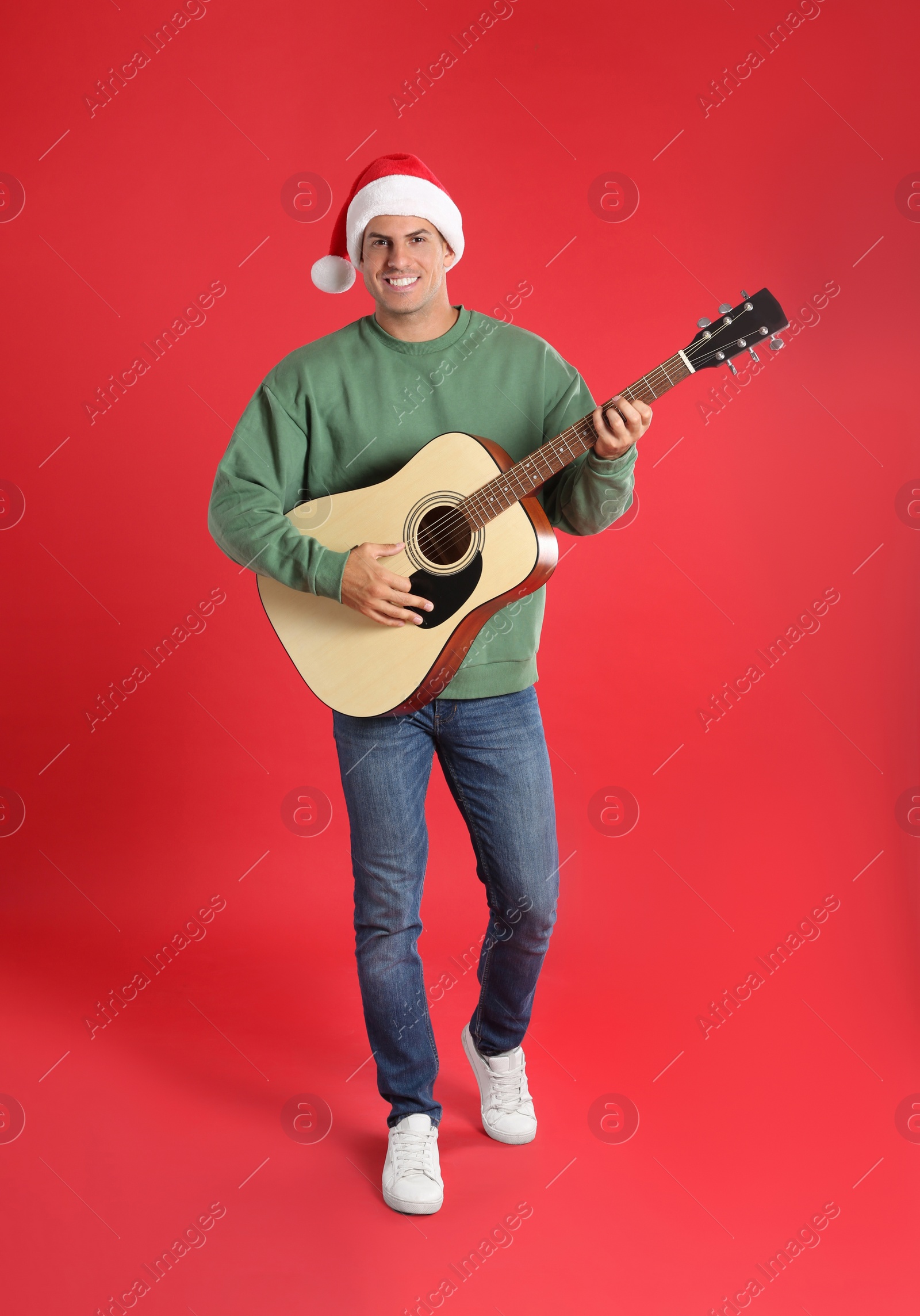 Photo of Man in Santa hat playing acoustic guitar on red background. Christmas music