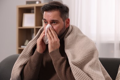 Photo of Sick man wrapped in blanket with tissue blowing nose on sofa at home. Cold symptoms