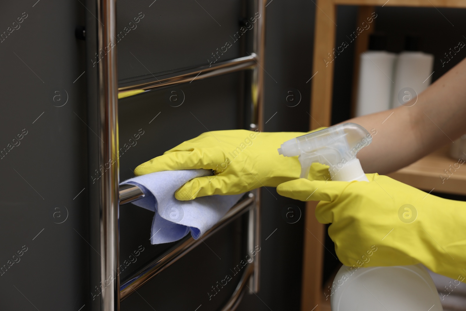 Photo of Woman cleaning heated towel rail with sprayer and rag indoors, closeup