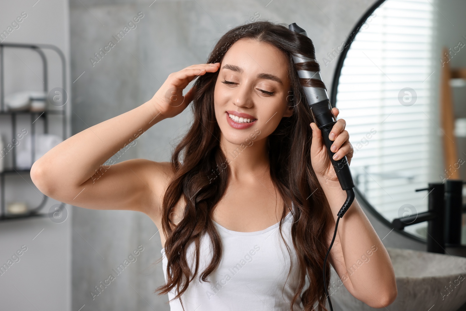 Photo of Smiling woman using curling hair iron in bathroom