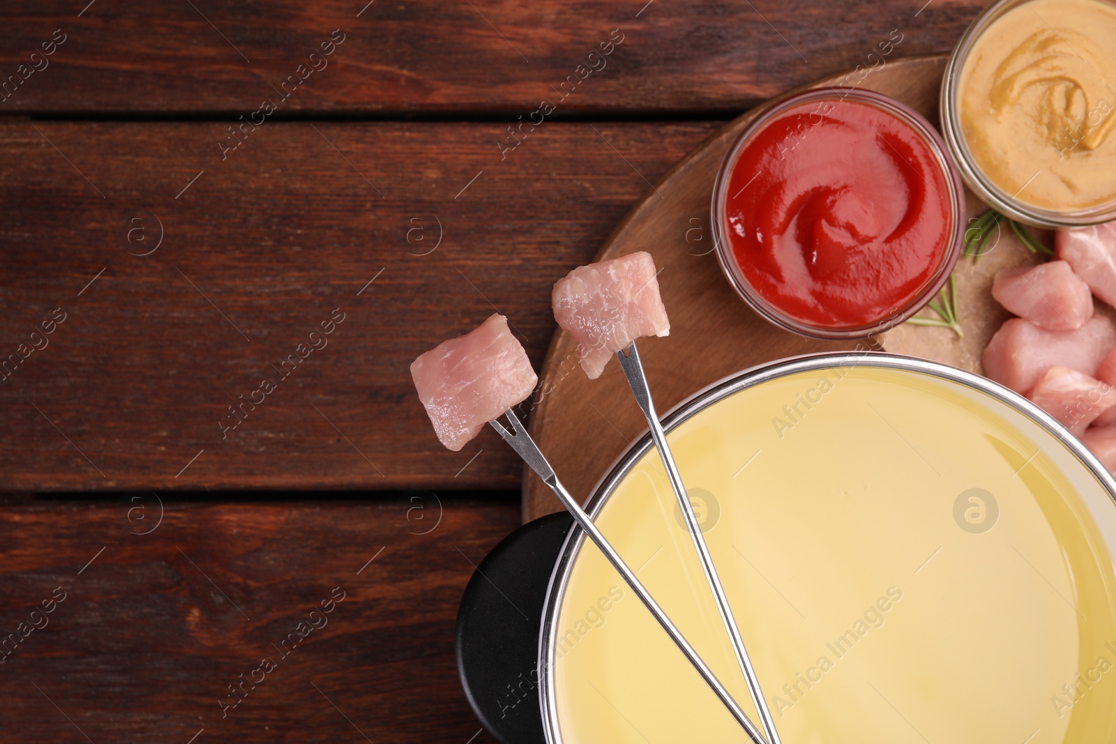 Photo of Fondue pot, forks with pieces of raw meat and sauces on wooden table, flat lay. Space for text