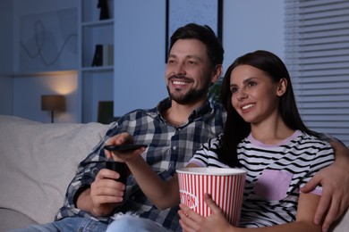 Photo of Happy couple watching show at home in evening. Woman holding popcorn and changing TV channels with remote control