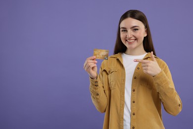 Happy woman pointing at credit card on purple background, space for text