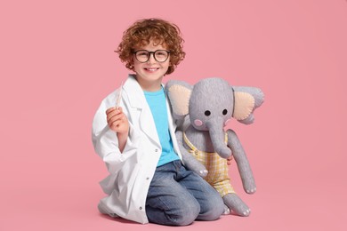 Photo of Little boy in medical uniform with thermometer and toy elephant on pink background