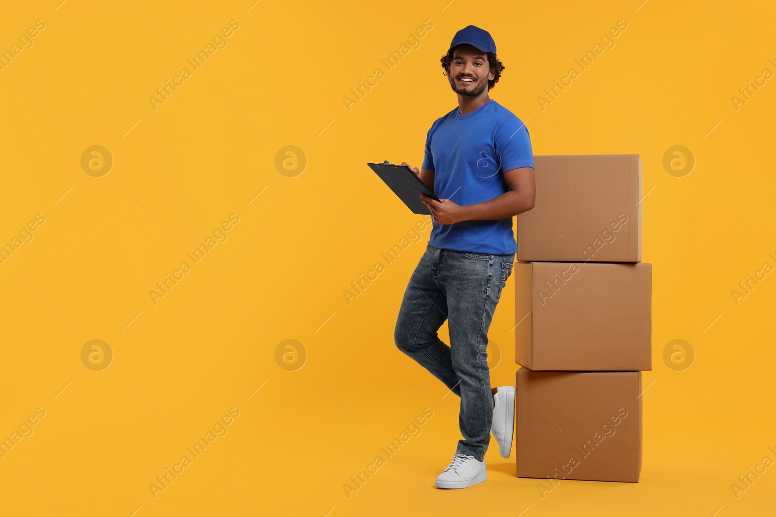 Photo of Happy courier with clipboard and stack of parcels on orange background, space for text