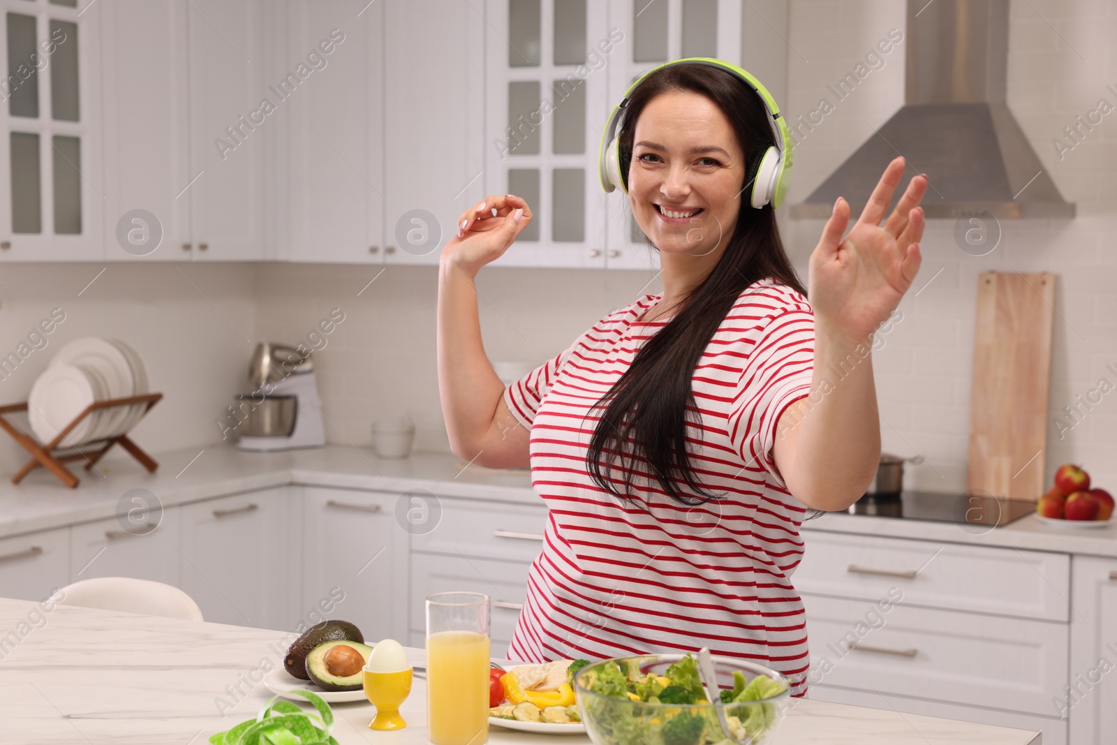 Photo of Happy overweight woman with headphones dancing near table in kitchen. Healthy diet