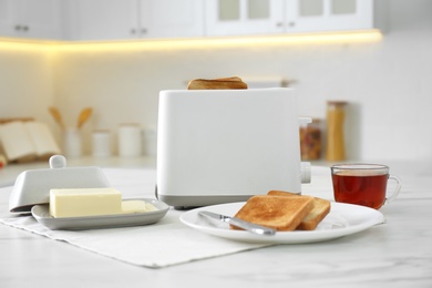 Photo of Modern toaster with bread slices, cup of tea and butter on white marble table in kitchen