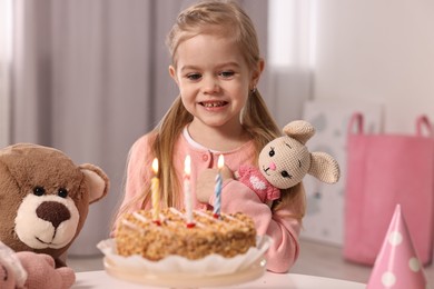 Cute girl with birthday cake and toys at table indoors