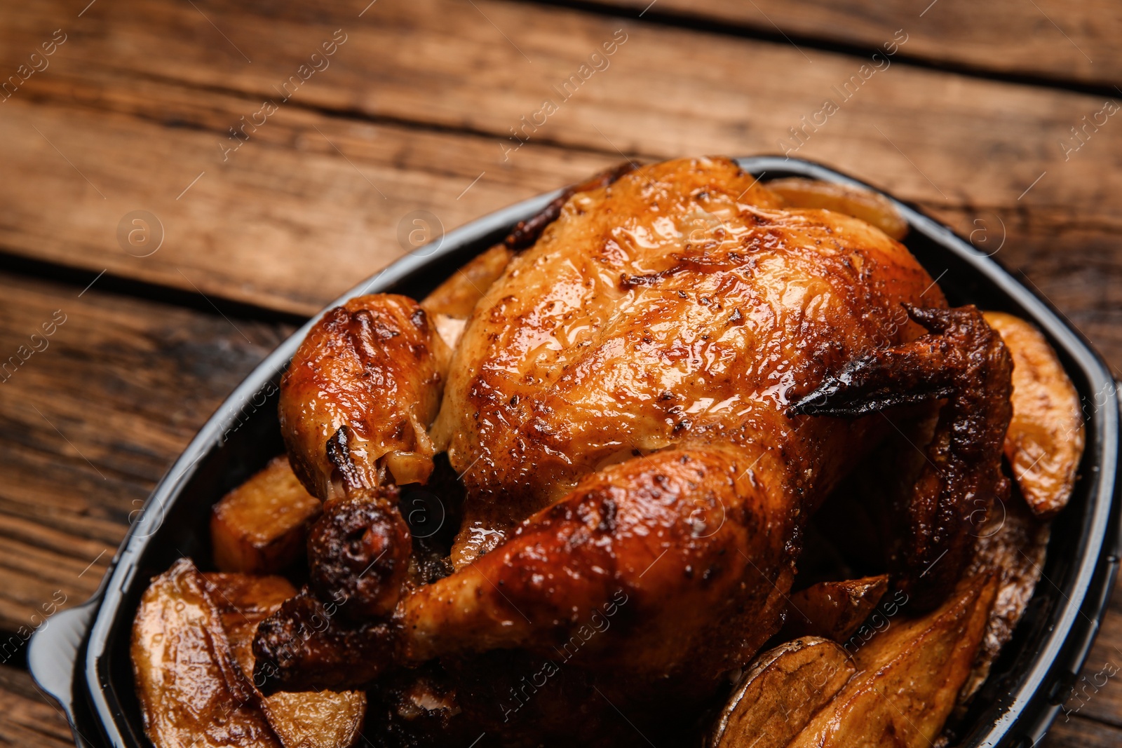 Photo of Delicious grilled whole chicken with potato in plastic container on wooden table, closeup. Food delivery service