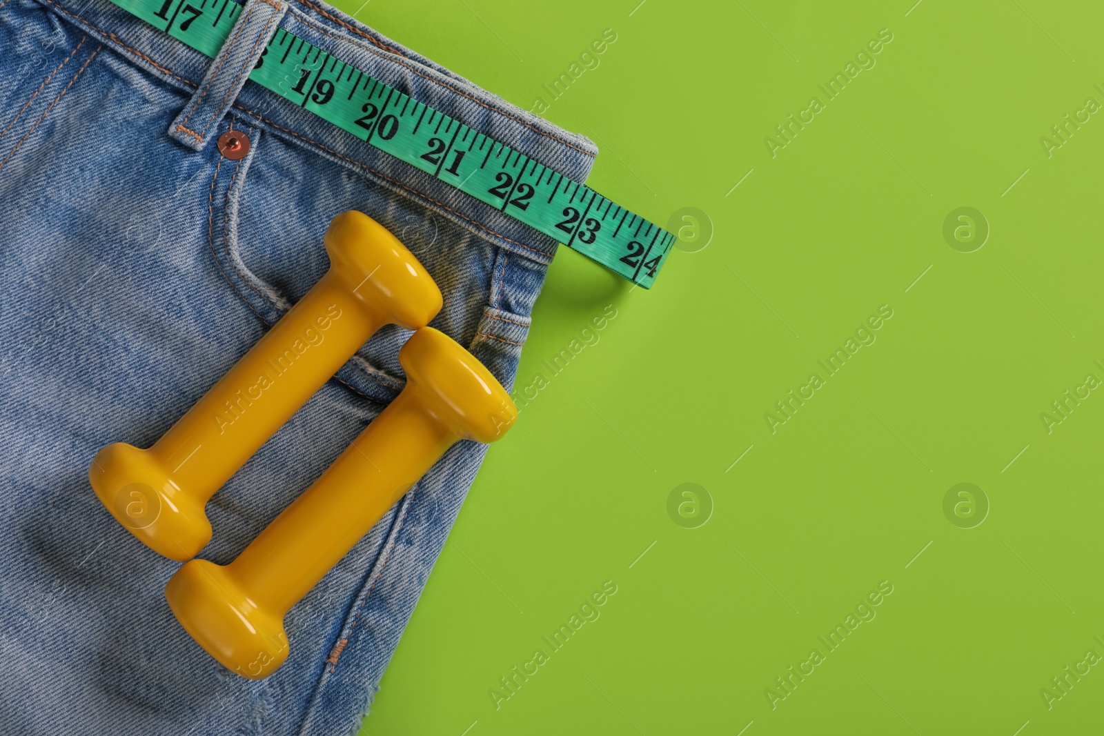 Photo of Jeans, dumbbells and measuring tape on light green background, flat lay with space for text. Weight loss concept