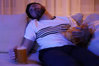 Photo of Man with chips, pizza and glass of beer sleeping on sofa at night. Bad habit