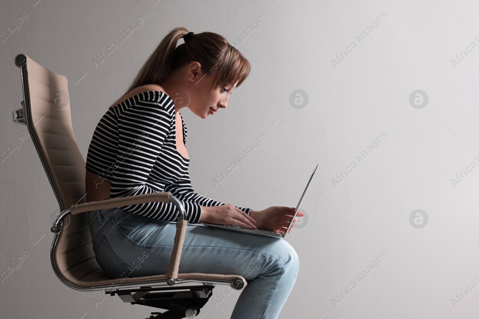 Photo of Young woman with poor posture using laptop while sitting on chair against grey background, space for text