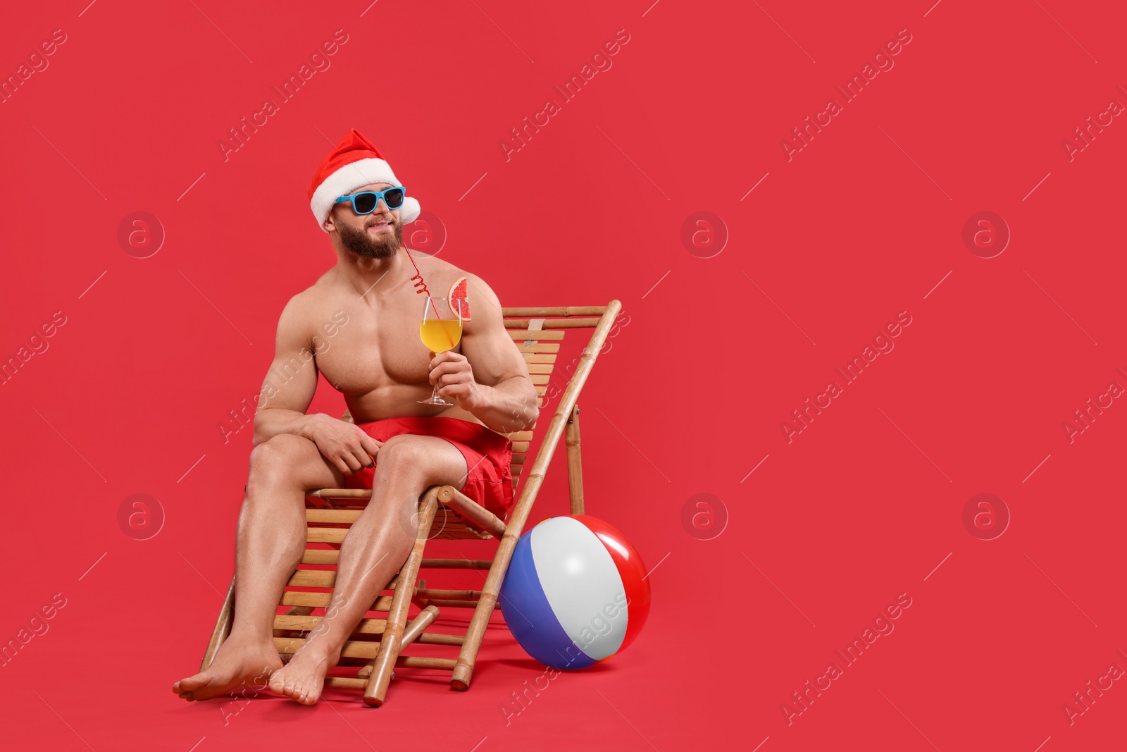 Photo of Muscular young man in Santa hat with cocktail, ball, sunglasses and deck chair on red background, space for text