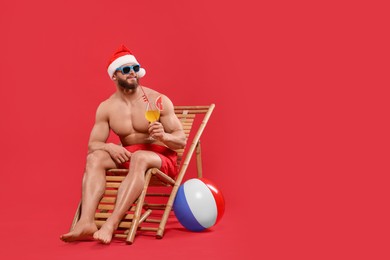 Muscular young man in Santa hat with cocktail, ball, sunglasses and deck chair on red background, space for text