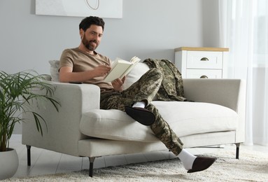 Photo of Happy soldier reading book on soft sofa in living room. Military service