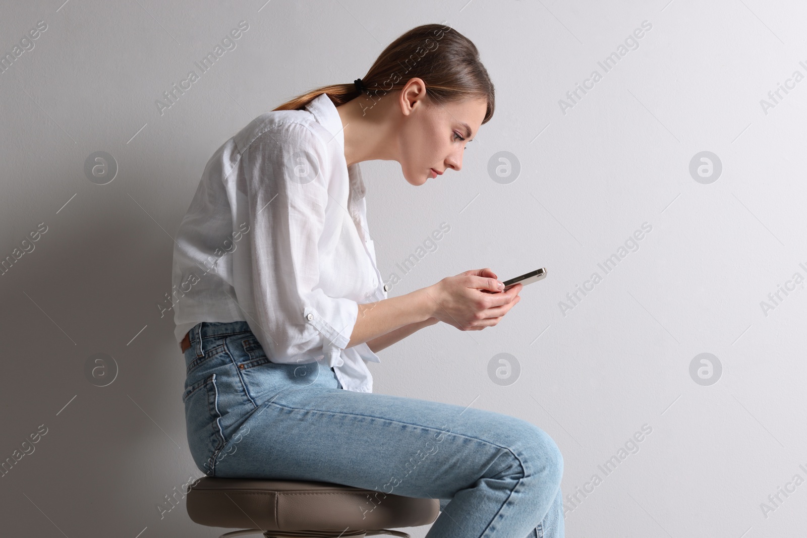 Photo of Woman with bad posture using smartphone while sitting on stool against light grey background