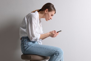 Photo of Woman with bad posture using smartphone while sitting on stool against light grey background