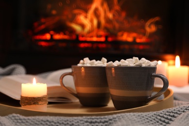 Photo of Cups of hot drink, candle and open book near fireplace indoors